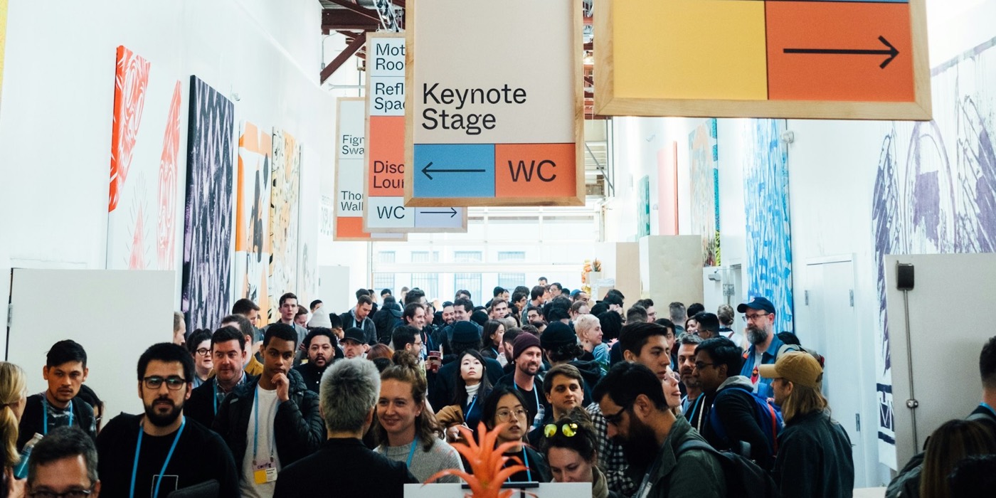 Attendees wearing lanyard badges fill a conference hall under hanging signs for the keynote stage and maker space.